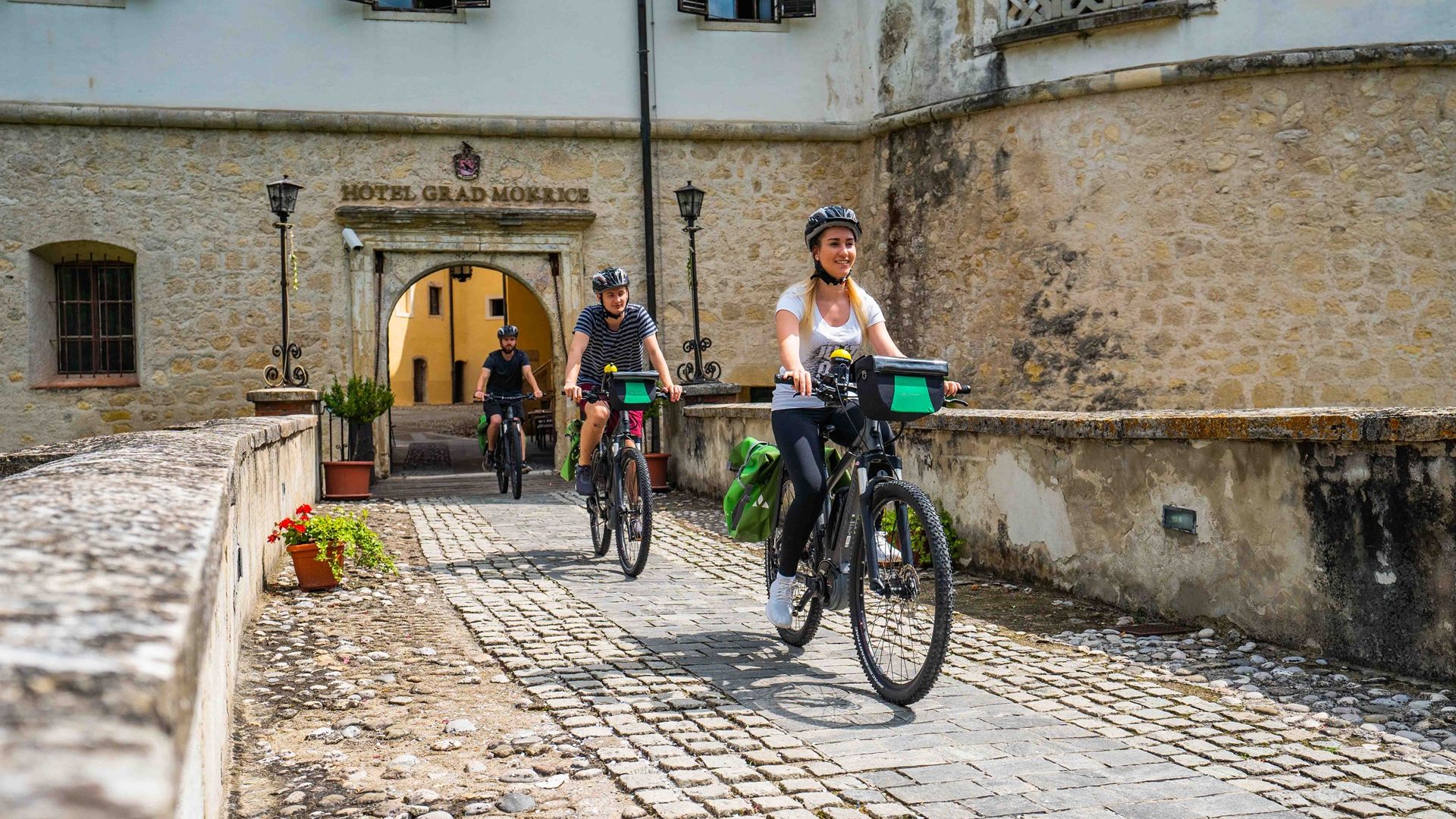 Trije kolesarji, ki kolesarijo po kamnitem mostu, zapuščajo hotel Grad Mokrice.