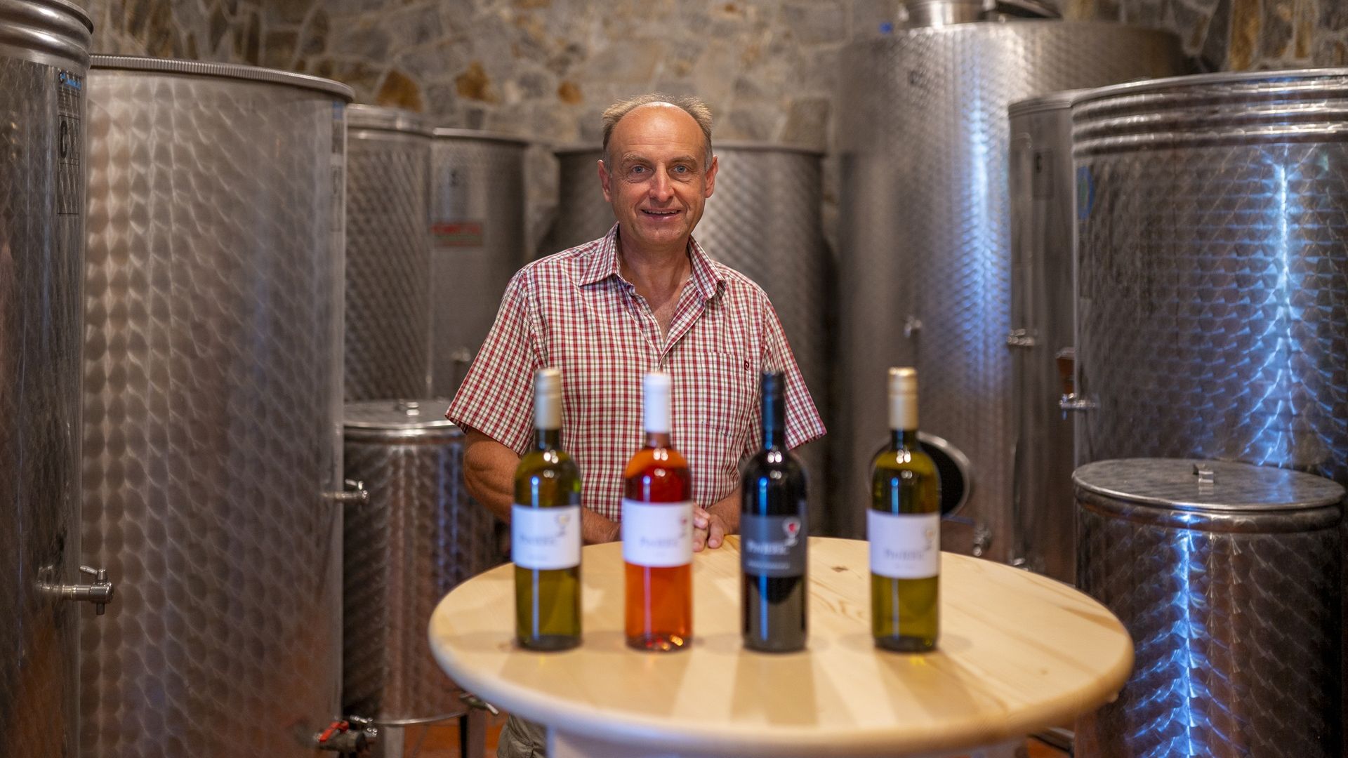 Fotografija moškega, ki stoji za mizo s štirimi vini. Za njim so vinski sodi.