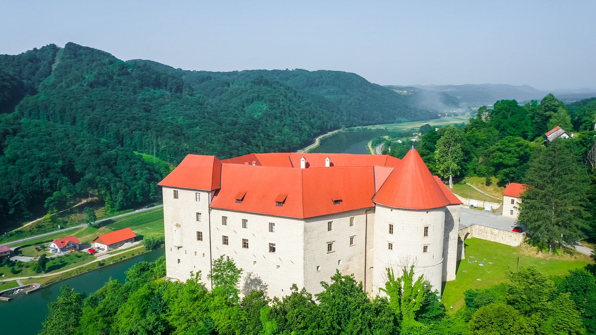 Fotografija gradu Rajhenburg, posneta iz zraka. Mimo njega teče reka, v ozadju pa se vzpenja gozdnat grič.