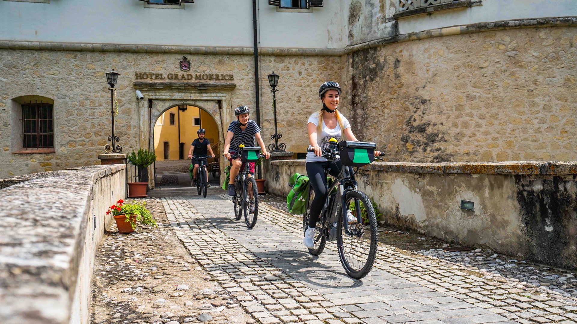 Trije kolesarji, ki kolesarijo po kamnitem mostu, zapuščajo hotel Grad Mokrice.